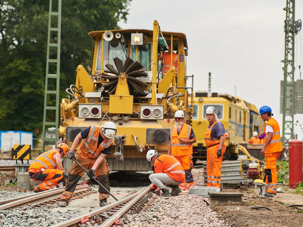 Arbeiter werkeln auf dem Gleis. Im Hintergrund eine Schotterplaniermaschine.