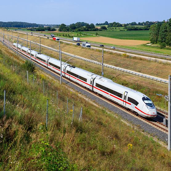 Ein ICE in Fahrt in einer grünen Landschaft, daneben ist eine befahrene Straße zu sehen