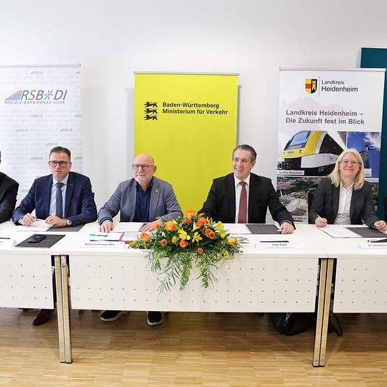 Fünf Männer und eine Frau sitzen an einem langen Tisch und lächeln in die Kamera