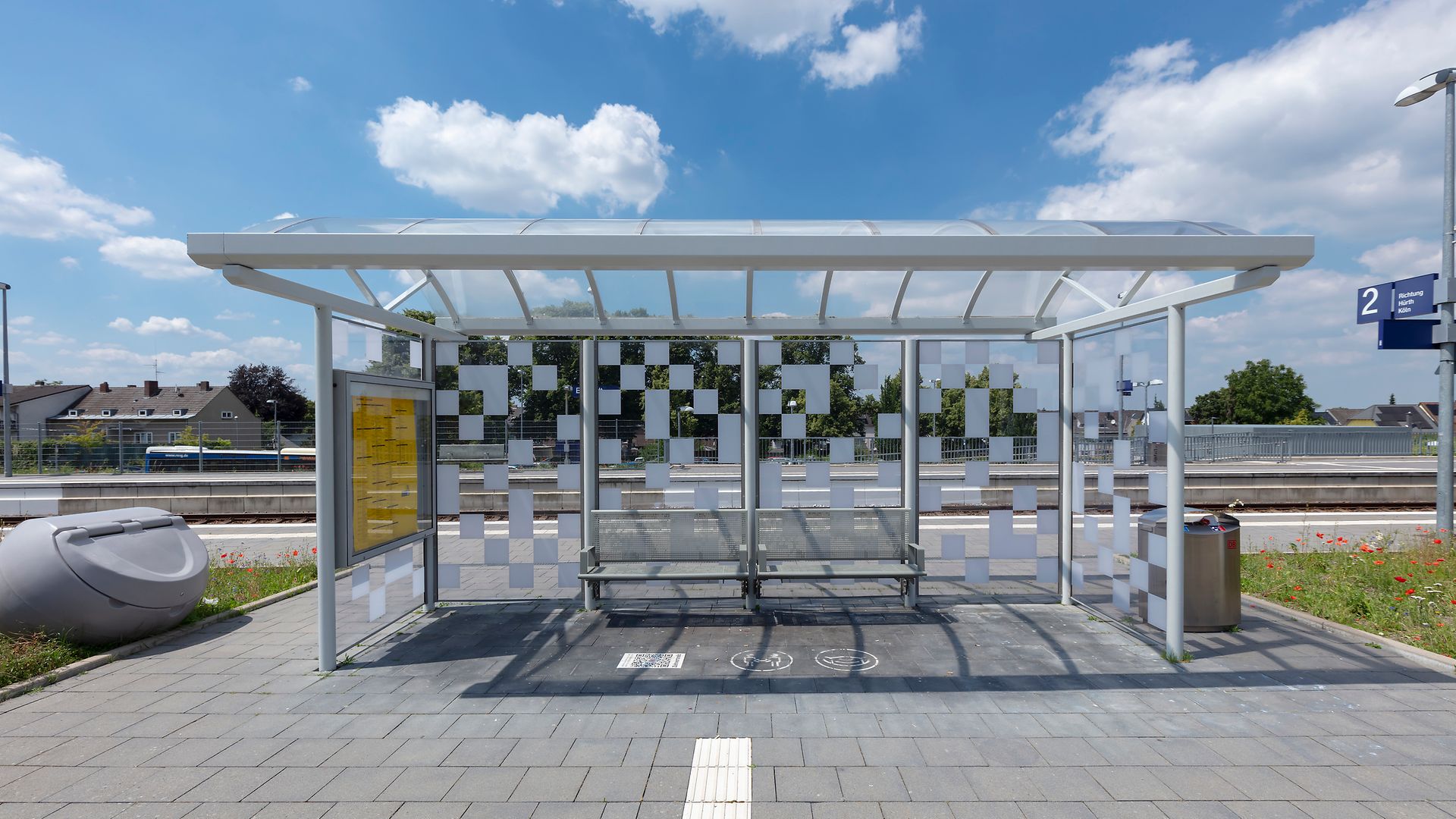 Ein gläsernes Wartehäuschen mit Sitzbänken am Bahnsteig unter blauem Himmel