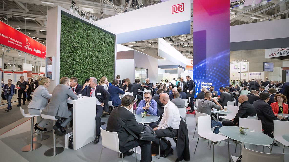 Messestand der Deutschen Bahn mit Pflanzenwand, DB-Logo und vielen Menschen, die an Tischen sitzen und sich austauschen.
