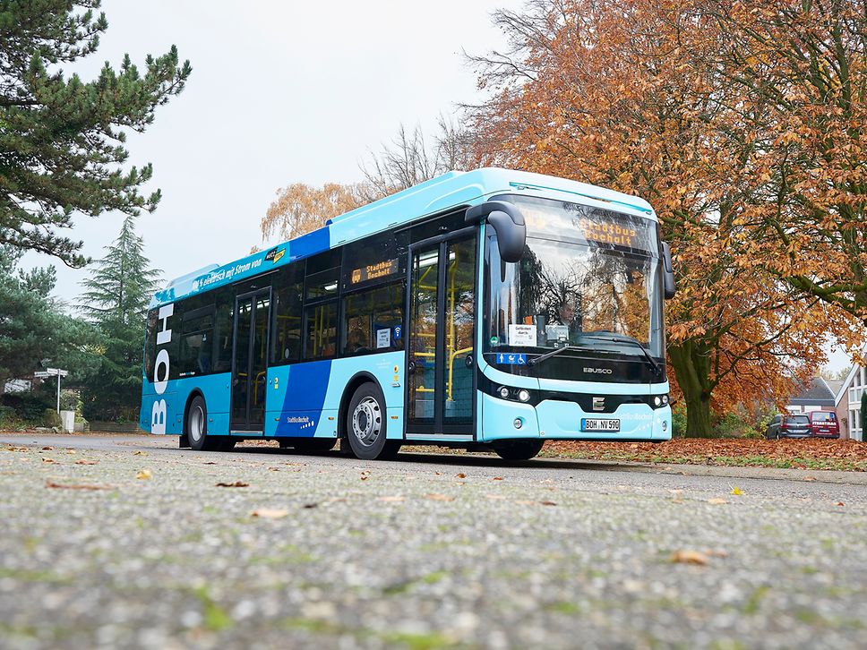 Ein hellblauer Stadtbus steht an einer Straße mit herbstlichen Bäumen im Hintergrund