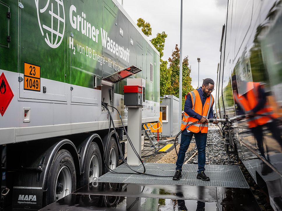 A man in a high-visibility vest stands between a tanker trailer and a train, holding a fuel hose in his hand.