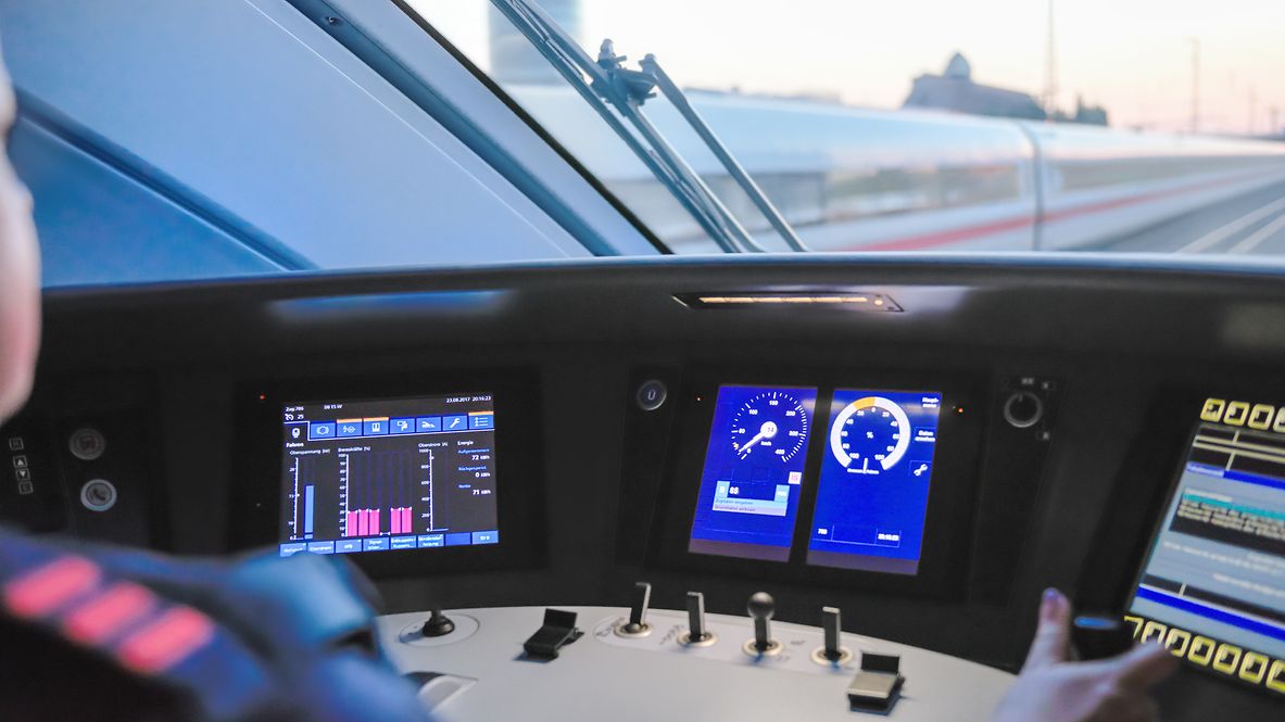 View of three different monitors in the driver's cab of an ICE train. A train driver can be seen dimly in the left-hand edge.